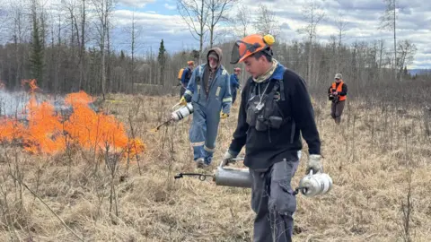 A man in an orange hard hat and black workwear carries gas cannister as a team behind him works on a controlled burn. There are four men behind him and they was through dry brush. There is a small fires to the side.