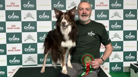 Mark Douglas Mark Douglas and his dog with the winning rosette, which is coloured red and green. They are sitting on a podium, in front of a Crufts advertising hoarding. Mark is wearing a dark green polo shirt and grey trousers. He is smiling and holding the dog on his lap. The dog is looking into the camera, it is alert and has its mouth slightly open.