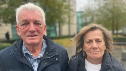 Mark and Majorie Sharp standing side by side outside. Mr Sharp has short grey hair and is looking at the camera as he wears a checked shirt and navy jacket. Mrs Sharp has shoulder-length brown hair and is wearing a white top and a black coat.