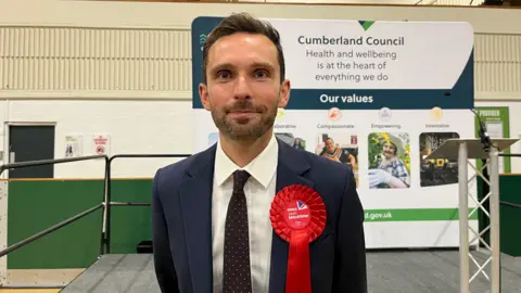 BBC Winning Labour candidate Josh McAlister wearing a blue suit and a red rosette