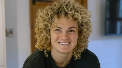 A woman with curly blonde hair smiles at the camera. She is wearing a black T-shirt.