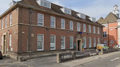 Google High Wycombe police station. A three story Geogian-style red brick building with tall white painted windows and more windows in the roof. A police van is sitting outside the building on the far right. 