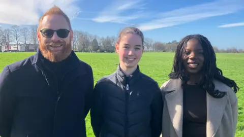 Greg, Alana and Adeolu stand for the photo. They're stood in a green field on a sunny day. Greg has ginger hair whilst Alana has her hair tied back. Adeolu has long black hair.
