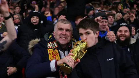 Scotland fans inside Hampden Park football stadium, with two fans holding a replica trophy in their hands - one of the fans is kissing it.