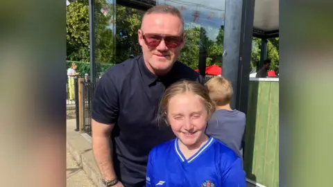 Paul Styles Wayne Rooney with a young football fan. He wears a black polo shirt and sunglasses and she wears a blue football top.