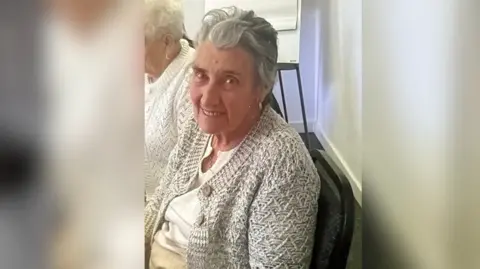Supplied Joyce Sinclair pictured from the side wearing a grey cardigan, beige shirt and a pendant around her neck. She has her head turned and is smiling at the camera.