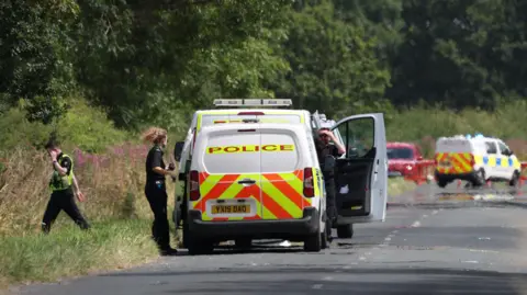 PA Media Three police vans parked on a road surrounded by trees and bushes. Two police officers at the door of the closest van and a police officer walking into the woodland.