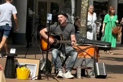 WES DOLAN A man sat on a chair with a guitar in his hand singing into a microphone