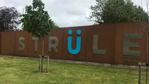 Signage for the Strule campus in Omagh, the wall is chocolate brown and the letters are a light brown colour except the U which is blue with two dots on top of it. The sky is grey and there are a couple of green trees, one in front of the wall and two behind the wall.