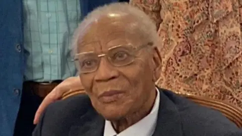 Shirley Henry/BBC An elderly man with clear rimmed glasses and white short hair is pursing his lips as he leans slightly forwards in a chair. Other people can be seen standing behind him. He is wearing a black blazer and white shirt.