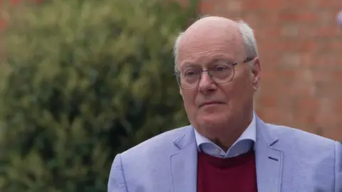 Sir John is sitting down in a garden and starring off camera at the interviewer. He is wearing a blue suit, burgundy jumper and blue shirt. 