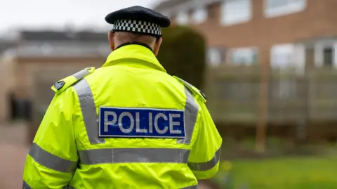 A policeman walking away from a home wearing a bright yellow jacket with police on the back and a black and white hat