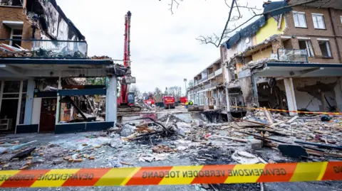 EPA A damage apartment block with a wide gap in the middle where the explosion took place. There is rubble an wood on the ground.  In the background is a large digger. In the foreground is a strip of emergency crew tape.