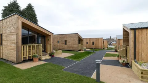 Frame PR A housing village - two rows of small wood-panelled homes, facing each other, with grass and turf to walk on in-between them.