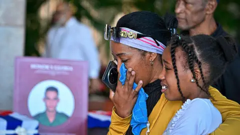 A woman wipes her tears while hugging a young girl, as a picture of a soldier can be seen in the background