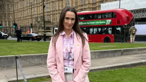 Monica Thomas A woman stands outside of Westminster in London. She has long brown hair and wears a pink blazer with a pink shirt and cream trousers. Vehicles pass along a road behind her. 