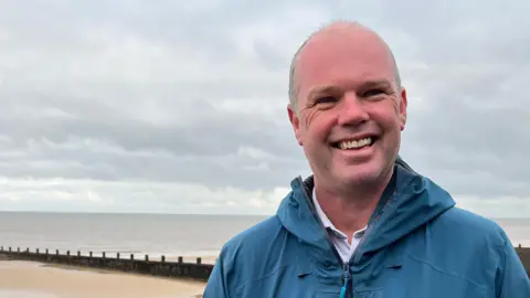 Clare Worden / BBC Andrew Raine stands on the stony beach at Sheringham. He is wearing a blue jacket and has short cropped greying hair. 