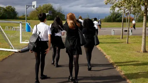 A group of secondary school students walk together across a school ground. They have their backs to the camera but they are wearing black uniforms with a white shirt. They are walking towards a tarmac playground surrounded by grassed areas, trees and sports equipment.