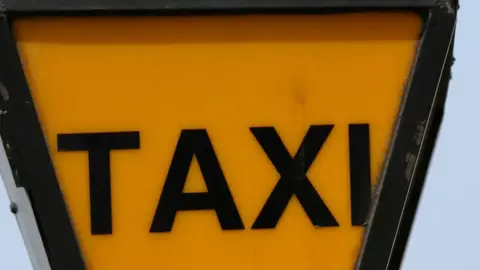BBC An orange lit sign with black writing reading TAXI.