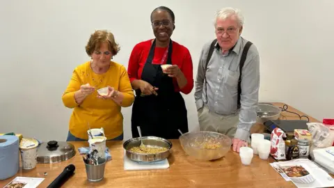 Hope Centre Three people, two women and a man stand behind a table where cooking instruction is being given. The women have small pots in their hands while the man has a large bowl in front of him on the table. Cutlery, pans and food ingredients are on the table.