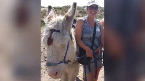 Louise Peeters Louise Peeters is a white woman wearing a blue and white striped vest and light coloured shorts, sunglasses and a cotton baseball-style cap. She has a black bag slung across her body. She is standing on the beach with a white donkey to her left holding a leading rein attached to a halter. She is smiling straight at the camera.