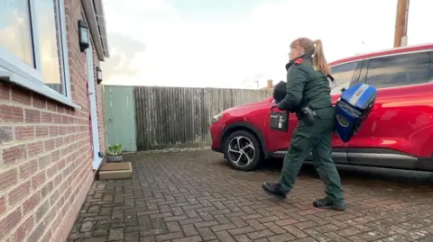 Chrissy Ames walks across a brick paved driveway towards the front door of a red brick house. She has a large blue holdall over her shoulder, a black bag under one arm and, in her hand, a device like an iPad in a hard plastic case. She is wearing the all green uniform of a paramedic and steel toecapped black boots. 