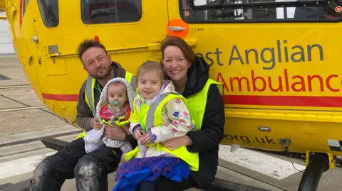 EAAA Russ Whitehouse, Claire Whitehouse, both in hi-vis waistcoats, sitting on the footboard of a yellow East Anglian Air Ambulance helicopter, while holding their young daughters on their laps 