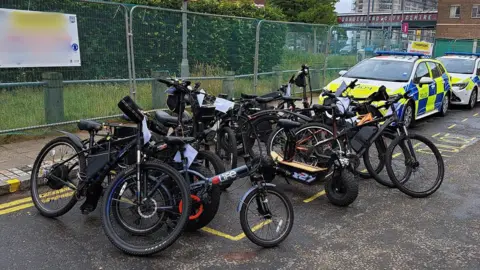 A number of seized e-bikes alongside two police cars.
