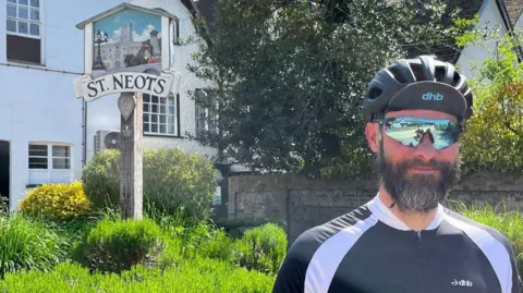 Richard Slade is standing in front of a grassy backdrop with the town sign of St Neots behind him. He is wearing a tight-fitting blue and white cycling top, a bicycle helmet and has sunglasses on. He has a greying beard and moustache.