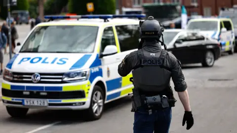 TT A police officer in tactical gear stands in front of a Swedish police car