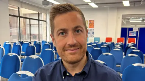 Head and shoulders image of Glasgow 2026 chief Phil Batty. He's sitting in a room full of blue chairs. He's looking directly at the camera and has spikey strawberry blond hair with a reddish goatie beard and moustache. He is wearing a navy blue shirt.