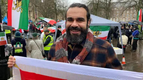 Kaveh Darafshi is standing on a square holding an Iranian flag and smiling at the camera. There are more people with flags in the background.