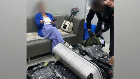 Thai Customs Department A woman sits on a sofa gesturing with one hand whilst holding a phone in another, her legs crossed, and her face is blurred. Meanwhile some in uniform is searching through a bag full of black vacuum compressed bags.