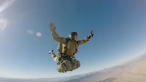 Collect/PA Real Life Rob during a parachute jump over the desert. His arms are stretched out, with his legs in the air behind him. He is wearing a khaki vest and a helmet with a clear visor, with what appears to be a kit bag attached to his harness. The ground below him is flat, with a mountain ridge in the distance. 