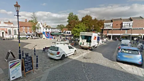 Hitchin Market Square, showing cars parked down one side and a lorry being unloaded. In the foreground are A-boards to shops and a lamppost. In the distance are shops and an inflatable slide on the square.