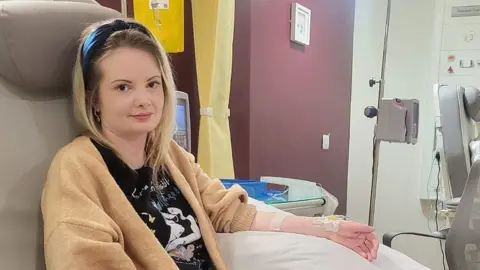 Gemma Coleman with long blond hair wearing a black headband and black t-shirt with a gold coloured cardigan sits in a medical room. Her sleeve on her left arm is rolled up and she has a drip going into her wrist. She is smiling.