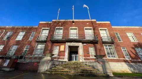 The Swindon Borough Council building seen from the front. It is a red-brick building with big windows. The sky behind it is blue.
