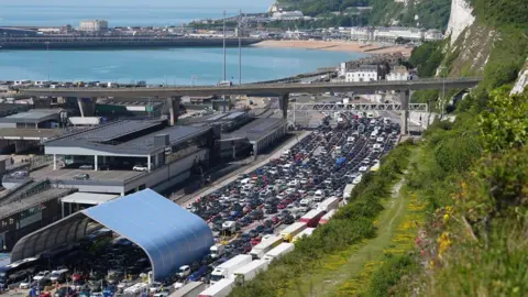 PA Media Traffic queues for the Port of Dover in Kent on 22 May 2026. Cars, vans and lorries queue in multiple lanes leading towards ferry and border control facilities. The port buildings sit beneath a road bridge, with the sea, harbour, beach and seafront visible in the background. White chalk cliffs and a grassy hillside run along the right-hand side of the image, overlooking the congested traffic.