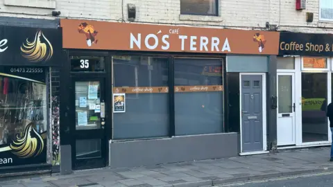 The frontage of Cafe Nos Terra in Ipswich. Its sign at the top of the building is orange with white writing.
