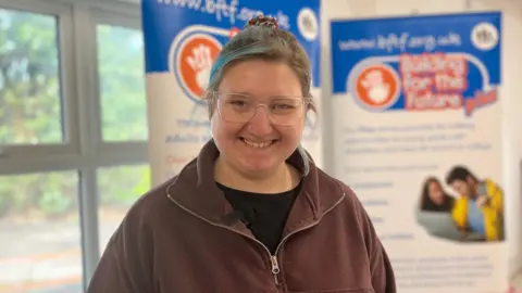 Hayleigh Woolven is wearing a brown zip-up jumper and is standing in front of a Building for the Future information board.