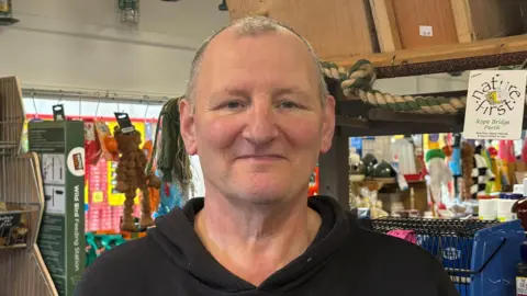 Man looking into camera, wearing black jumper and standing in pet food shop.