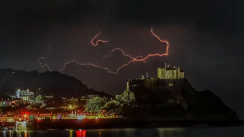 Andrew Battersby A red lightning bolt in the dark night sky above an illuminated old castle. Lights and buildings are also in the picture. 