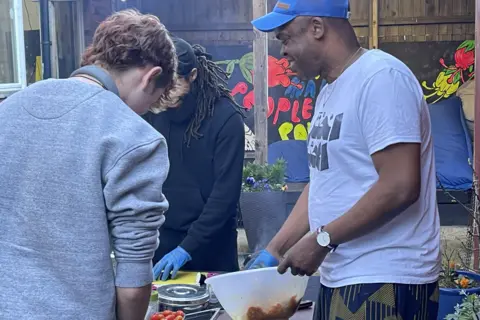 Oyindamola Odelabu Three people stand round a table. Brian Powlett is wearing a blue cap and one blue rubber glove and is holding a bowl. The other two people are chopping. 