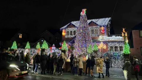 Paul Molyneux The image shows a two-story house completely covered in dazzling Christmas lights and decorations. A towering tree wrapped in multicolored lights stands in the front yard, surrounded by smaller illuminated trees and glowing ornaments. A crowd of people gathers on the pavement, admiring the festive display, while a car passes by in the foreground.
