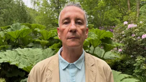 Tom Howell is pictured in the Vallee Des Vaux peace garden. He is wearing a beige coat and light blue shirt. 
