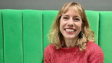 Hannah Shead has shoulder-length hair and is wearing a pink knitted jumper. She is sitting on a green seat and smiling broadly at the camera.