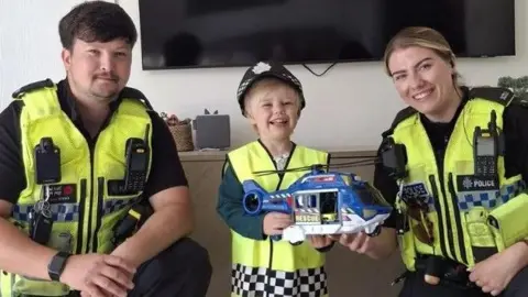 South Yorkshire Police A male police officer, PC Jordan Kassim, with brown hair and a beard wearing a police jacket, crouches next to a small blonde boy holding a toy police helicopter and wearing police hat and jacket. On the right is PC Charlotte Wright, a blonde woman wearing a yellow police jacket.