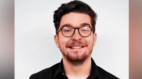 Supplied John Edwards is standing in front of a white background and looking directly into the camera while smiling. He has short brown hair and brown glasses, and is wearing a black shirt.