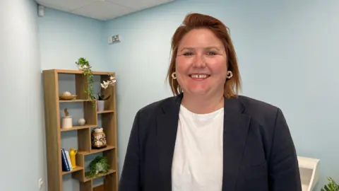 Jenny Murphy has short auburn hair and is wearing a white T-shirt and a navy suit jacket, smiling at the camera.
