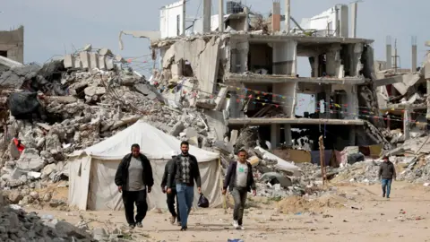 Palestinians walk among rubble in Rafah, with a tent and a bombed out building in the background (March 2025)